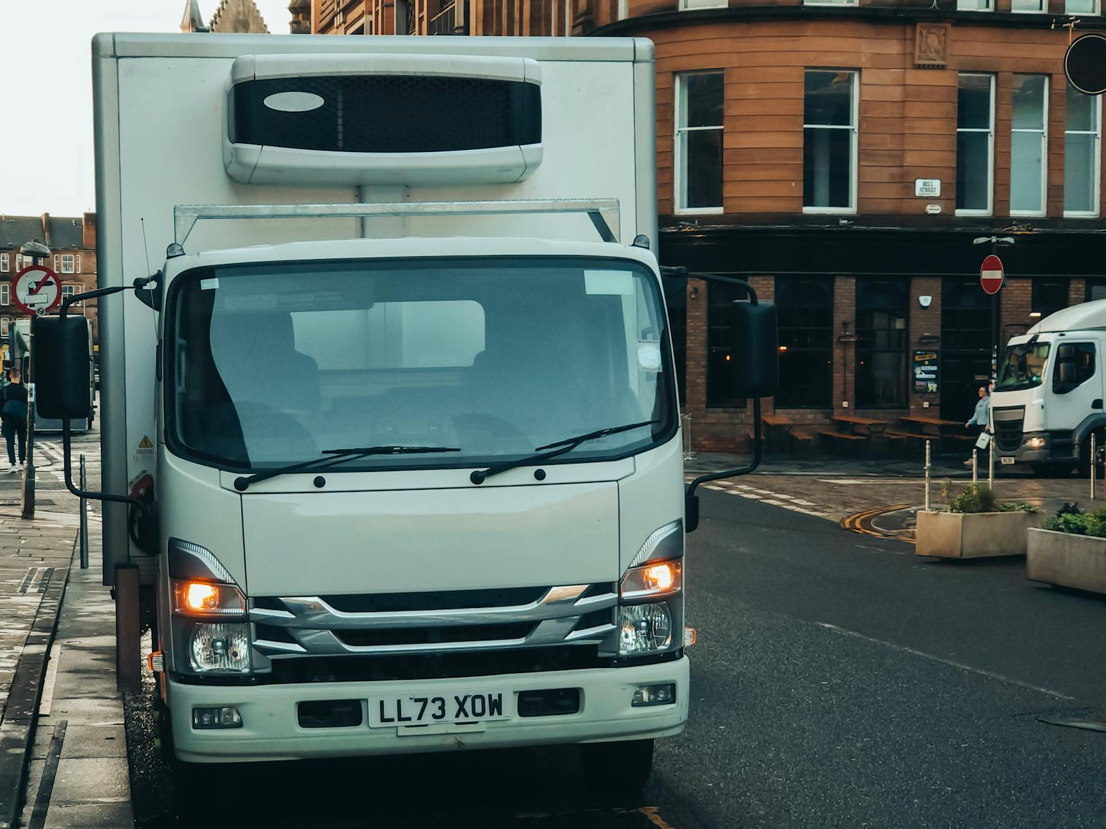 A white delivery truck parked on a city street with urban buildings in the background.